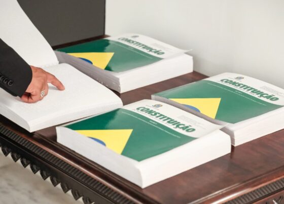 Foto em paisagem de cinco livros da Constituição Brasileira em braille com uma pessoa lendo
