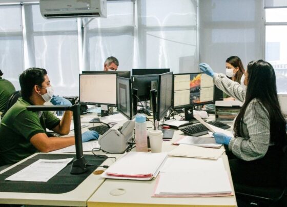 Foto em paisagem de um escritório com três homens utilizando máscara e luvas e duas mulheres usando máscaras e luvas. Todos trabalhando.