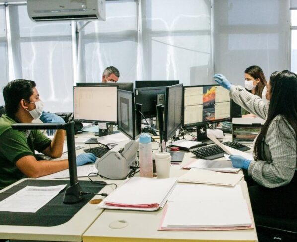 Foto em paisagem de um escritório com três homens utilizando máscara e luvas e duas mulheres usando máscaras e luvas. Todos trabalhando.