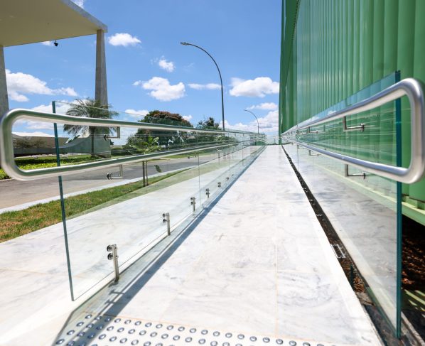 Imagem colorida horizontal de uma rampa com um corrimão cinza e proteção de vidro. Ao fundo o edifício-sede do STF e o céu azul