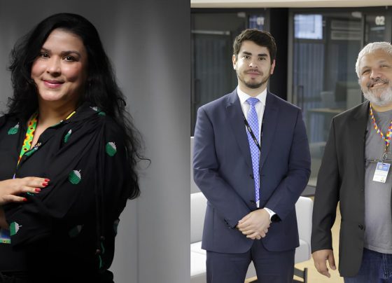 Foto colorida em formato horizontal. Á esquerda aparece uma mulher branca de cabelos pretos e cacheado, com o braço cruzado, roupas pretas e o cordão colorido com símbolos de quebra cabeça e a direita um homem adulto jovem com cabelos pretos e terno azul, e ao lado dele um homem com cabelos grisalhos e barba, usando blusa cinza, blaser preto e calça preta