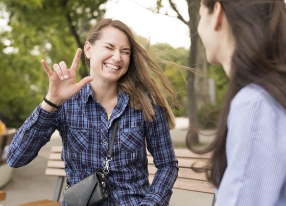 Duas mulheres estão sentadas em um banco ao ar livre. Uma delas sorri e faz o sinal de