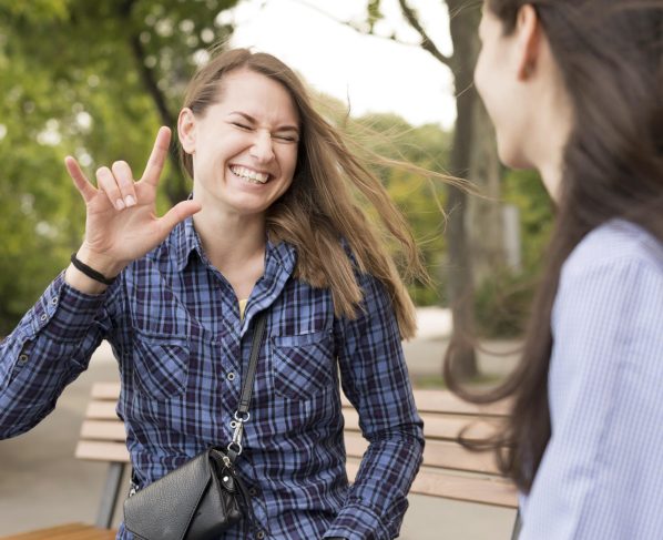 Duas mulheres estão sentadas em um banco ao ar livre. Uma delas sorri e faz o sinal de