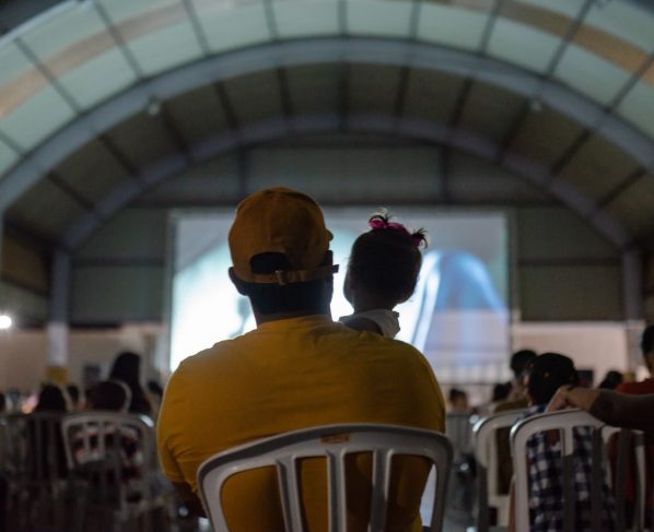 Governo de Goiás leva Cine Leitura do Bem a mais quatro municípios - público assistindo a sessão de cinema do projeto. Em primeiro plano, pai com bebê nos braços assistindo ao filme